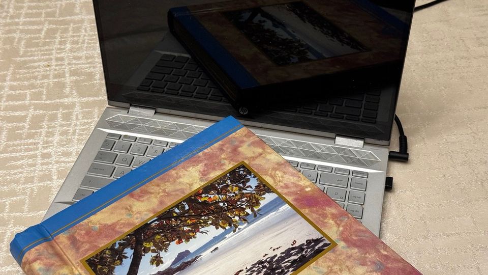 A hardcover photo album with a beach and a leafy tree on its cover, resting on the keyboard of an open laptop