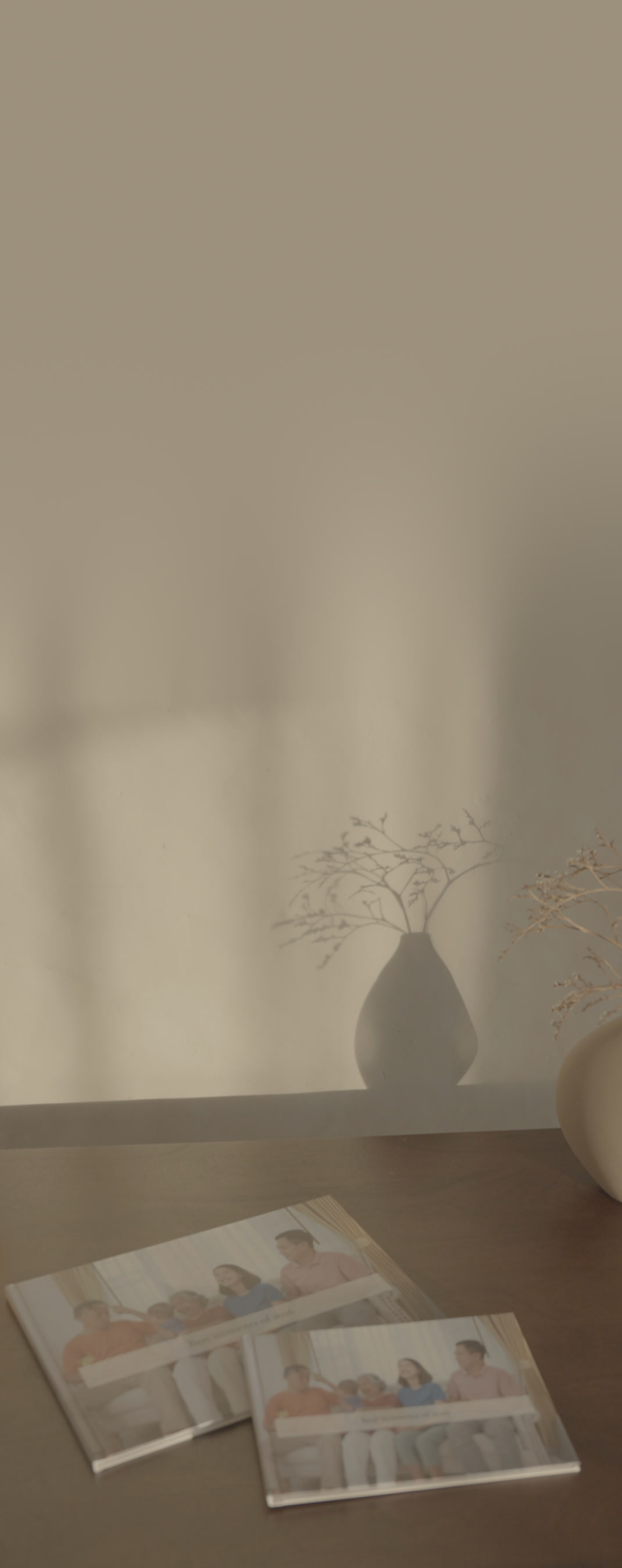 An A4 and an A5 Nokoru hardcover side by side on a dark wooden table below a sunlit wall and a vase of dried branches, the same family photo on both covers